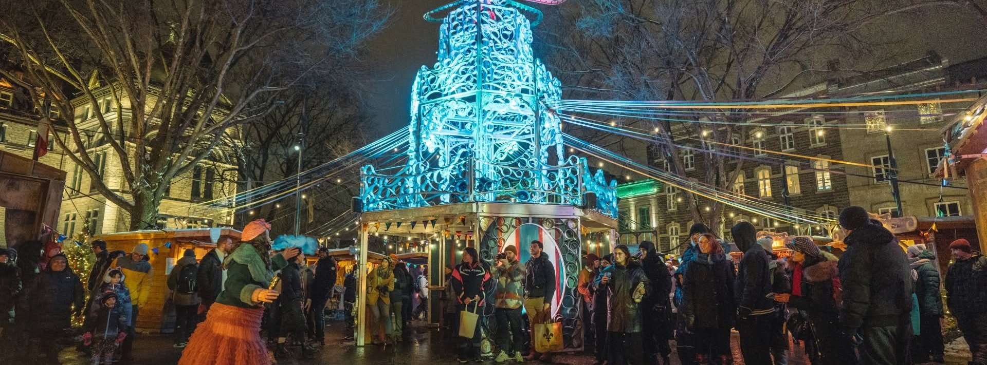 Festival Kaléidoscopes dans le Vieux-Québec : pyramide de Noël illuminée en bleu, artiste costumée du théâtre forain et foule en manteaux d’hiver devant les chalets, un soir de fin décembre
